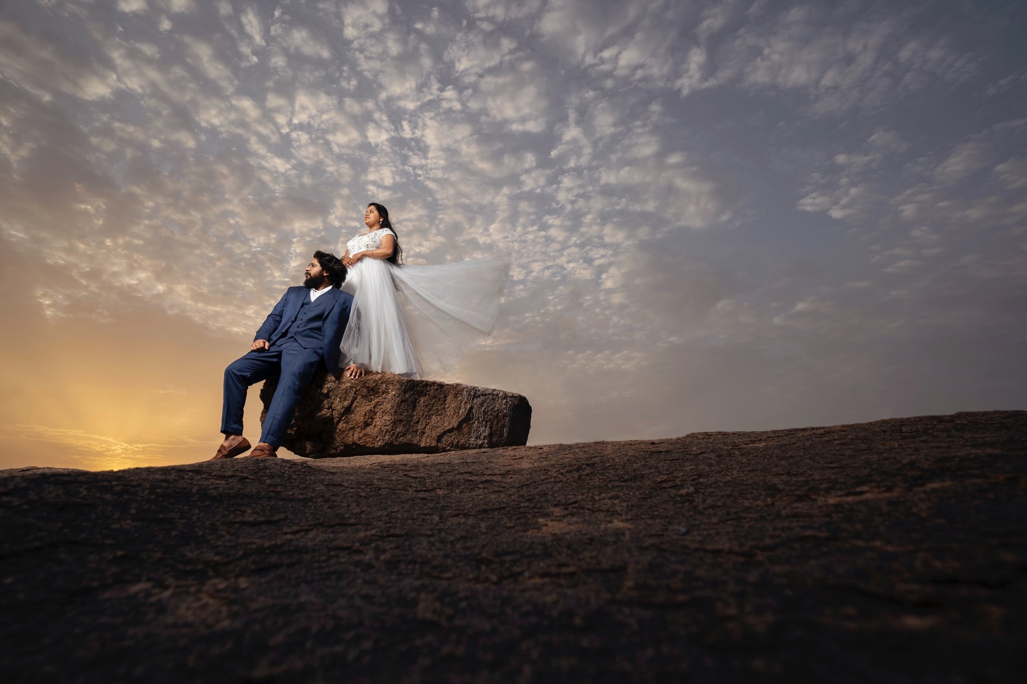 Cinematic wedding portrait of a couple on a rock formation against a dramatic sunset sky - perfect for wedding blogs and social media