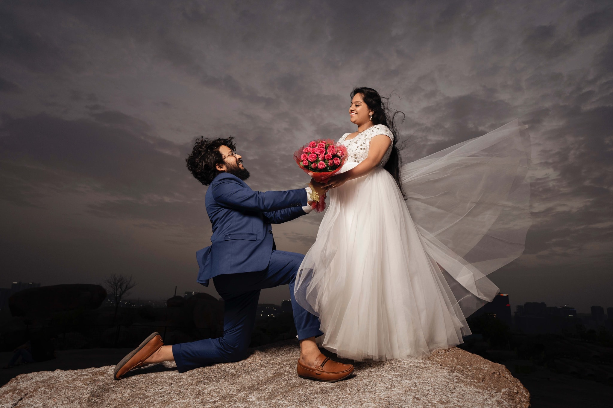 Cinematic wedding moment with groom kneeling to present roses to bride against dramatic sky backdrop