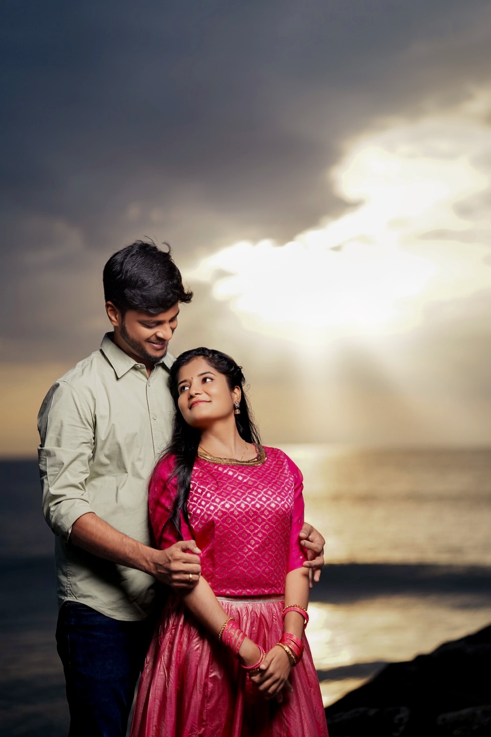 Romantic couple portrait at sunset by the ocean, the woman in a vibrant pink outfit and the man in light green, perfectly framed with dramatic golden light